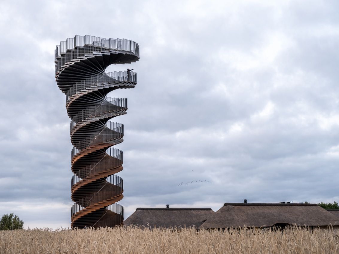 A tower in the middle of a wheat field. The tower is made of rusted corten steel, shaped like a double helix with two staircases.